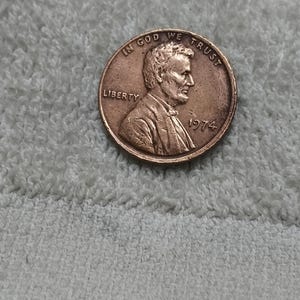 May include: A copper-coloured United States one-cent coin, commonly known as a penny, featuring the profile of Abraham Lincoln facing right. The coin is inscribed with the words IN GOD WE TRUST, LIBERTY, and the production year 1974.