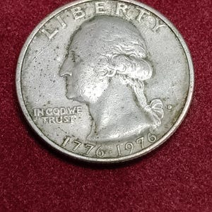 Op de afbeelding: Een zilverkleurige Amerikaanse kwart dollar munt met een profiel van George Washington. De tekst LIBERTY staat bovenaan, IN GOD WE TRUST aan de linkerzijde en de jaartallen 1776-1976 onderaan met een kleine D-muntteken.