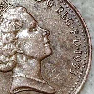 Peut inclure: Une pièce de monnaie en métal de couleur cuivre présentant un portrait de profil de la reine Elizabeth II tourné vers la droite. Le texte D.G.REG.F.D.1993 est gravé sur le bord droit. La pièce a une bordure perlée et une finition métallique.