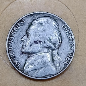 May include: A silver-colored United States five-cent coin featuring a profile of Thomas Jefferson facing left. The text IN GOD WE TRUST is printed on the left side, and the word LIBERTY and the year 1964 are printed on the right side of the coin.