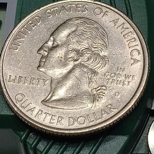 Op de afbeelding: Een zilverkleurige Amerikaanse kwart dollar munt met het profiel van George Washington naar links gericht. De munt bevat de tekst UNITED STATES OF AMERICA, LIBERTY, IN GOD WE TRUST en QUARTER DOLLAR, met een zichtbaar D-muntteken.