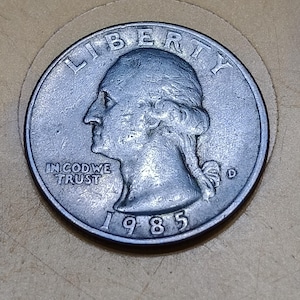 May include: A 1985 United States quarter dollar coin featuring the profile of George Washington facing left. The coin includes the text LIBERTY at the top, IN GOD WE TRUST on the left, the year 1985 at the bottom, and a D mint mark on the right.