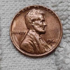 以下が含まれることがあります： 右を向いたエイブラハム・リンカーンの横顔が描かれた銅色の米国1セント硬貨。硬貨の上部には「IN GOD WE TRUST」、左側には「LIBERTY」、右側には「1968」という年号と「D」のミントマークが刻印されています。