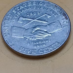 May include: A silver-colored United States five cents coin featuring a handshake and crossed axes. The text reads UNITED STATES OF AMERICA, LOUISIANA PURCHASE, 1803, E PLURIBUS UNUM, and FIVE CENTS. The coin has a circular shape and metallic finish.