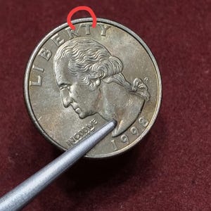 May include: A silver-colored 1998 United States quarter coin featuring the profile of George Washington. The word LIBERTY is arched at the top, with the phrase IN GOD WE TRUST and the year 1998 visible. A red circular mark highlights the letter T.