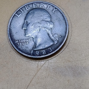 May include: A silver-colored United States quarter dollar coin dated 1983. The obverse side features a profile portrait of George Washington facing left, with the word LIBERTY above, the phrase IN GOD WE TRUST to the left, and the year 1983 below.