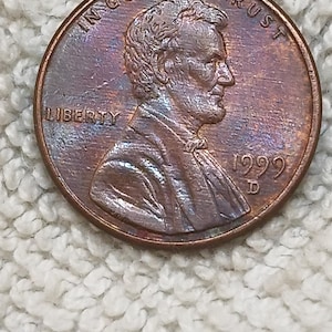 Op de afbeelding: Een koperkleurige Amerikaanse munt van één cent met het profiel van Abraham Lincoln naar rechts gericht. De munt is voorzien van de tekst IN GOD WE TRUST, LIBERTY en het jaartal 1999 met een D-muntteken onder de datum.