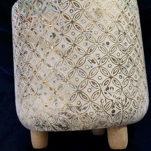 Può includere: Un vaso cilindrico in ceramica con base bianca e un motivo geometrico dorato anticato composto da cerchi e quadrifogli intrecciati. Il contenitore è sostenuto da tre piccoli piedini in legno di colore chiaro alla base.