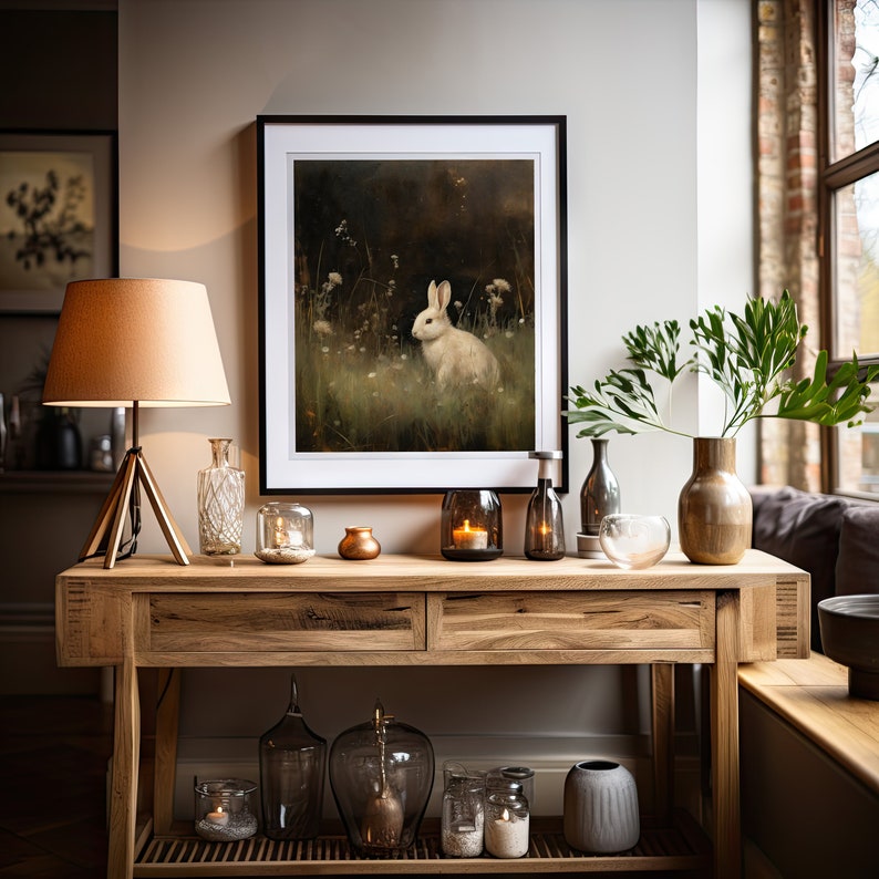May include: A wooden console table with two drawers and a lower shelf. The table is decorated with a framed print of a white rabbit in a forest, a lamp, vases, candles, and other decorative items.