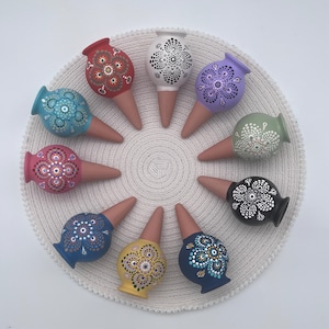Puede incluir: Nueve picos de riego de terracota dispuestos en círculo. Cada pico presenta una parte superior bulbosa pintada en un color sólido único, incluyendo rojo, blanco, lavanda, verde salvia, negro, azul oscuro, amarillo, azul medio y rosa con patrones de puntos.