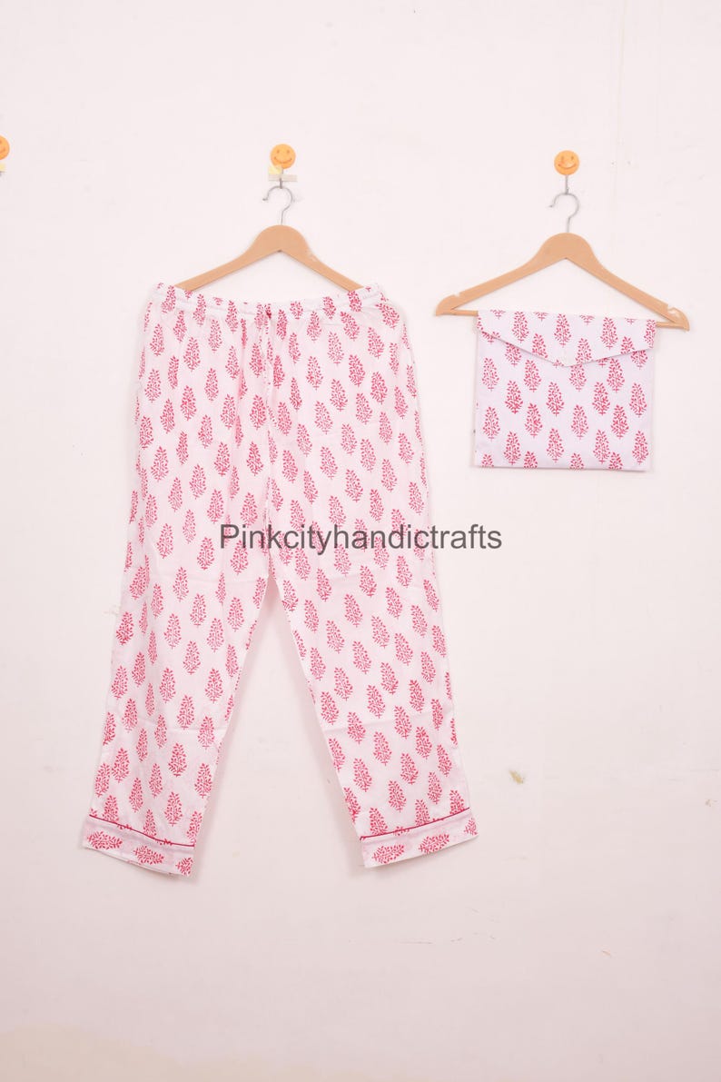 May include: White pajama set with a repeating pink floral pattern. The set includes trousers and a matching pouch, all hanging on wooden hangers. The text "Pinkcityhandicrafts" is visible on the trousers.