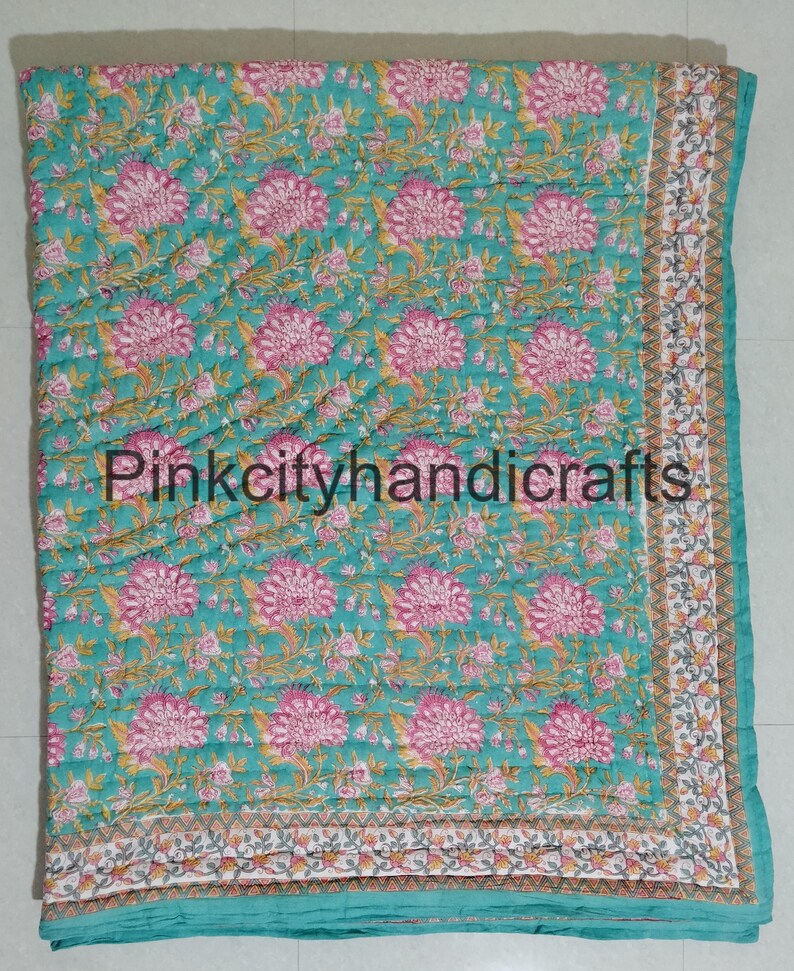 Puede incluir: Una colcha de algod&oacute;n azul turquesa con un patr&oacute;n floral en tonos de rosa y verde. La colcha tiene un borde blanco con un patr&oacute;n geom&eacute;trico gris y blanco.