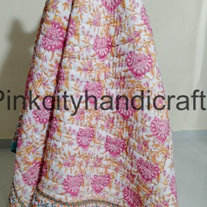 Puede incluir: Una colcha de algod&oacute;n blanco con estampado floral en tonos rosa y amarillo. La colcha tiene un borde turquesa con un patr&oacute;n geom&eacute;trico. La colcha est&aacute; doblada y drapeada sobre una superficie.