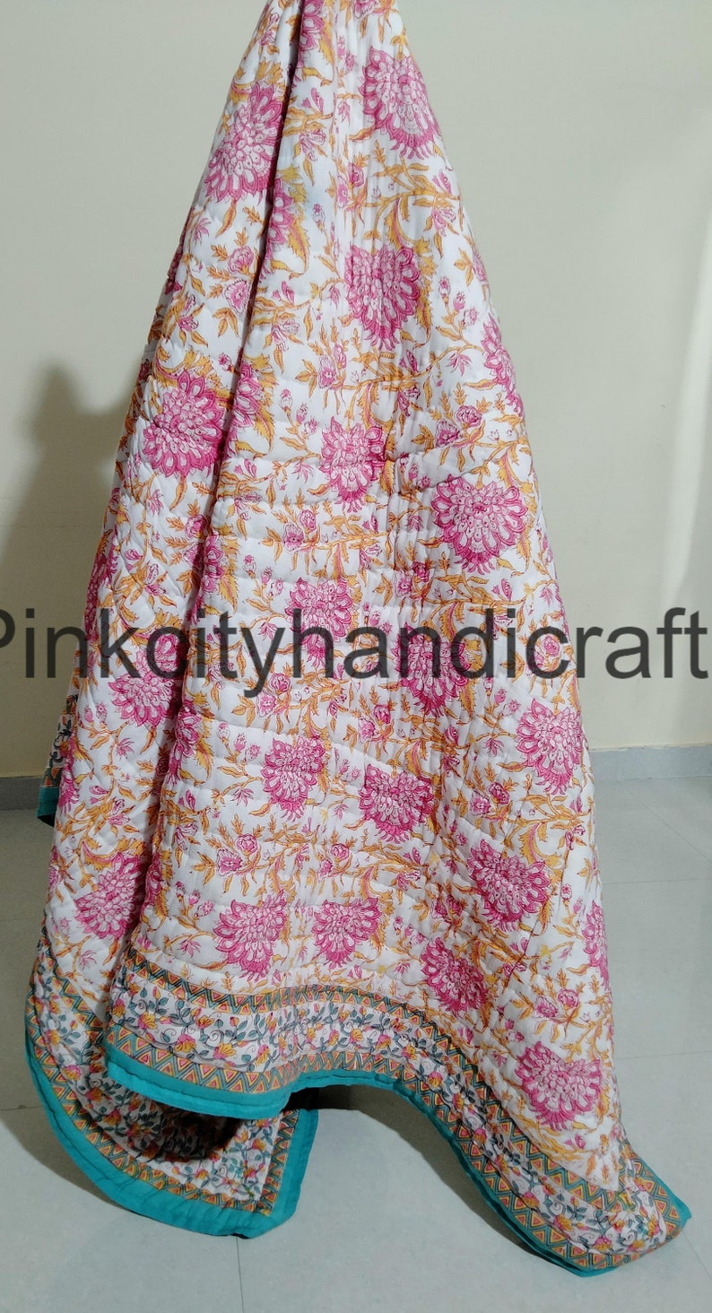 Puede incluir: Una colcha de algod&oacute;n blanco con estampado floral en tonos rosa y amarillo. La colcha tiene un borde turquesa con un patr&oacute;n geom&eacute;trico. La colcha est&aacute; doblada y drapeada sobre una superficie.