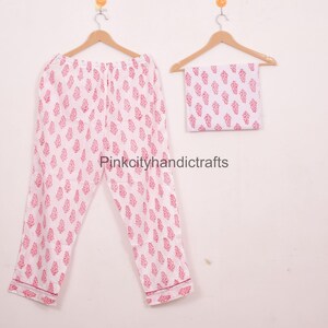 May include: A white pajama set with a pink leaf pattern, including trousers and a matching square piece of fabric, hangs on wooden hangers. The pajama trousers are folded and have a pink trim. The set is displayed against a plain white background.