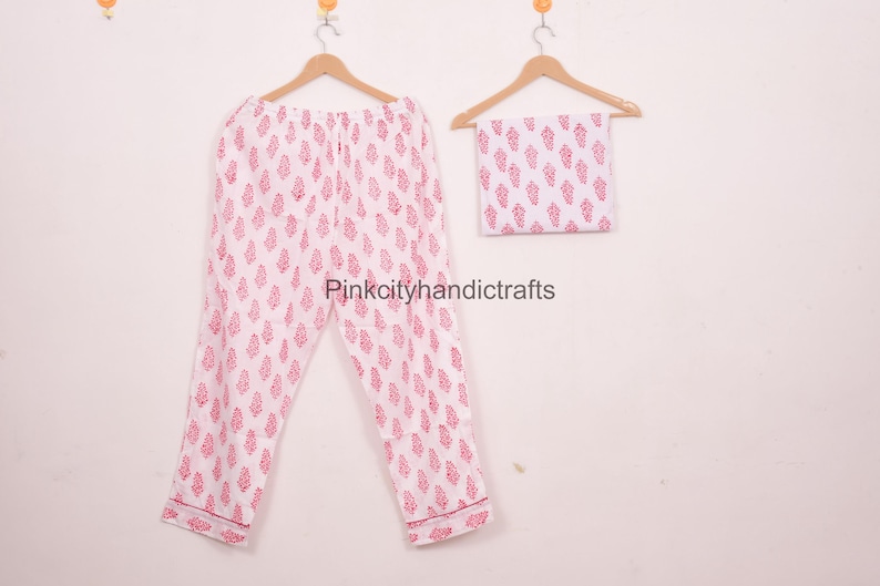May include: A white pajama set with a pink leaf pattern, including trousers and a matching square piece of fabric, hangs on wooden hangers. The pajama trousers are folded and have a pink trim. The set is displayed against a plain white background.