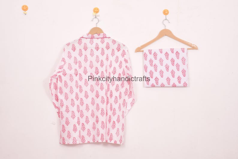 May include: White pajama set with a red floral pattern, including a long-sleeved top and matching folded bottoms, hanging on wooden hangers. The top has a collared neckline. The text "Pinkcityhandicrafts" is visible.