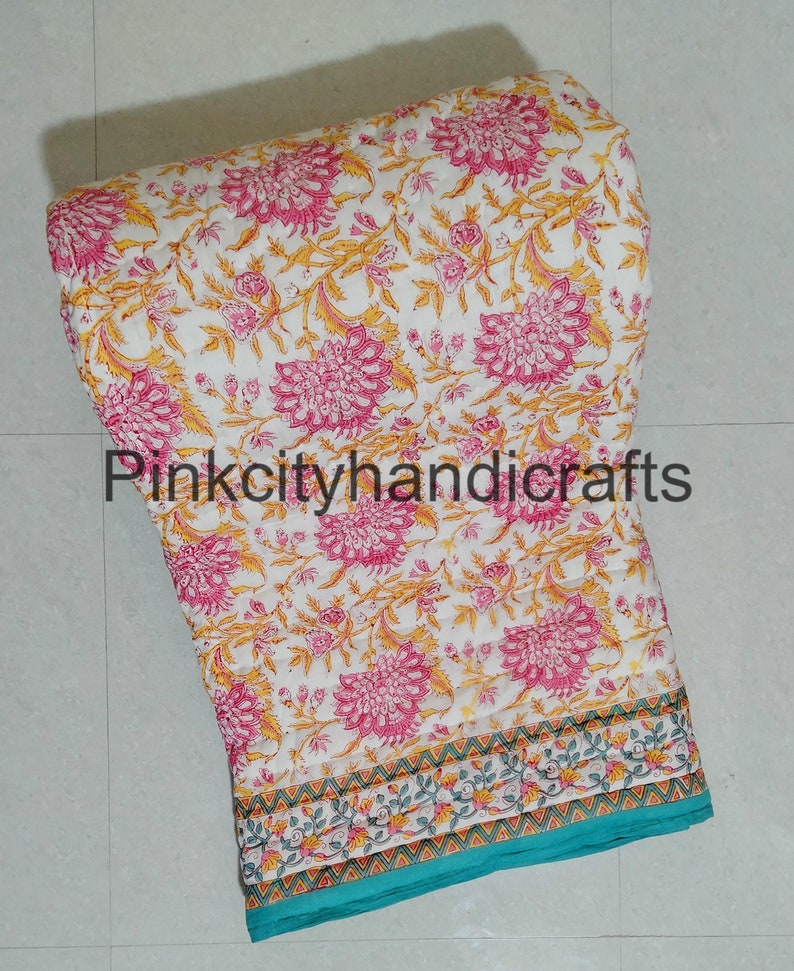 Puede incluir: Una colcha de algod&oacute;n blanco con estampado floral en tonos rosa y amarillo. La colcha tiene un borde turquesa con un patr&oacute;n floral blanco y amarillo. La colcha est&aacute; doblada y enrollada, y el texto "Pinkcityhandicrafts" es visible en el centro de la imagen.
