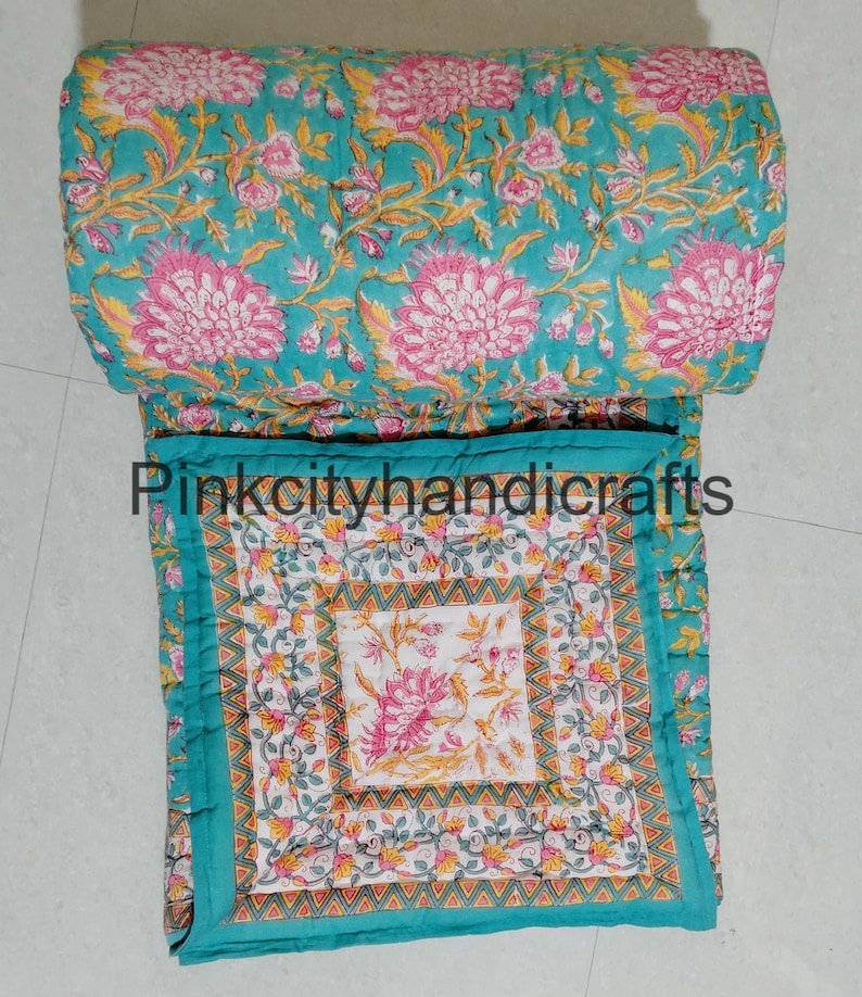 Puede incluir: Una colcha de algod&oacute;n azul turquesa con estampado floral. La colcha est&aacute; doblada por la mitad y el estampado floral es visible en ambos lados. La colcha est&aacute; cosida a mano y tiene un dise&ntilde;o tradicional indio.