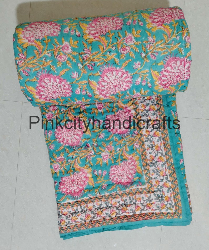 Puede incluir: Una colcha de algod&oacute;n azul turquesa con estampado floral en tonos rosa, amarillo y verde. La colcha tiene un borde con un patr&oacute;n geom&eacute;trico en blanco, gris y naranja.