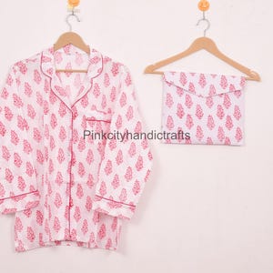 May include: White pyjama set with a red floral pattern, including a button-down shirt and matching pouch. The shirt has a notched collar and red trim. The set is hanging on a wooden hanger.