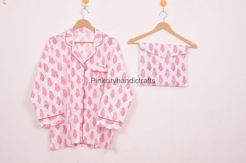 May include: White pyjama set with a red floral pattern, including a button-down shirt and matching pouch. The shirt has a notched collar and red trim. The set is hanging on a wooden hanger.