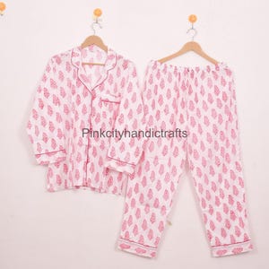 May include: White pyjama set with a pink floral pattern. The long-sleeved top has a button-down front and a notched collar. The trousers have a drawstring waist. The set is hanging on a wooden hanger.