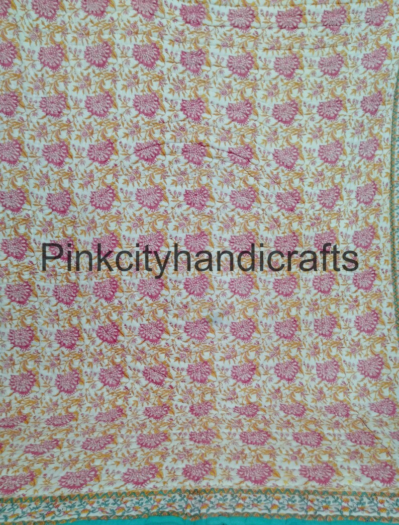 Puede incluir: Una colcha de algod&oacute;n blanco con un patr&oacute;n floral en tonos rosa y amarillo. La colcha tiene un borde turquesa con un ribete blanco y amarillo.