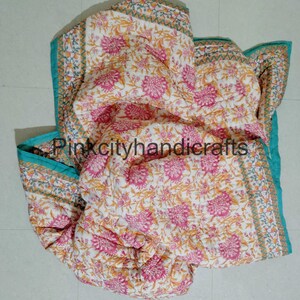 Puede incluir: Una colcha de algod&oacute;n blanco con estampado floral en tonos rosa, amarillo y verde. La colcha tiene un borde turquesa con un patr&oacute;n geom&eacute;trico. La colcha est&aacute; doblada y colocada sobre un suelo de baldosas blancas.