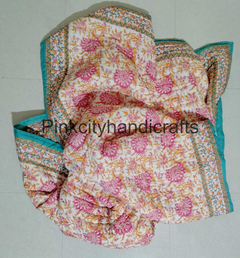 Puede incluir: Una colcha de algod&oacute;n blanco con estampado floral en tonos rosa, amarillo y verde. La colcha tiene un borde turquesa con un patr&oacute;n geom&eacute;trico. La colcha est&aacute; doblada y colocada sobre un suelo de baldosas blancas.