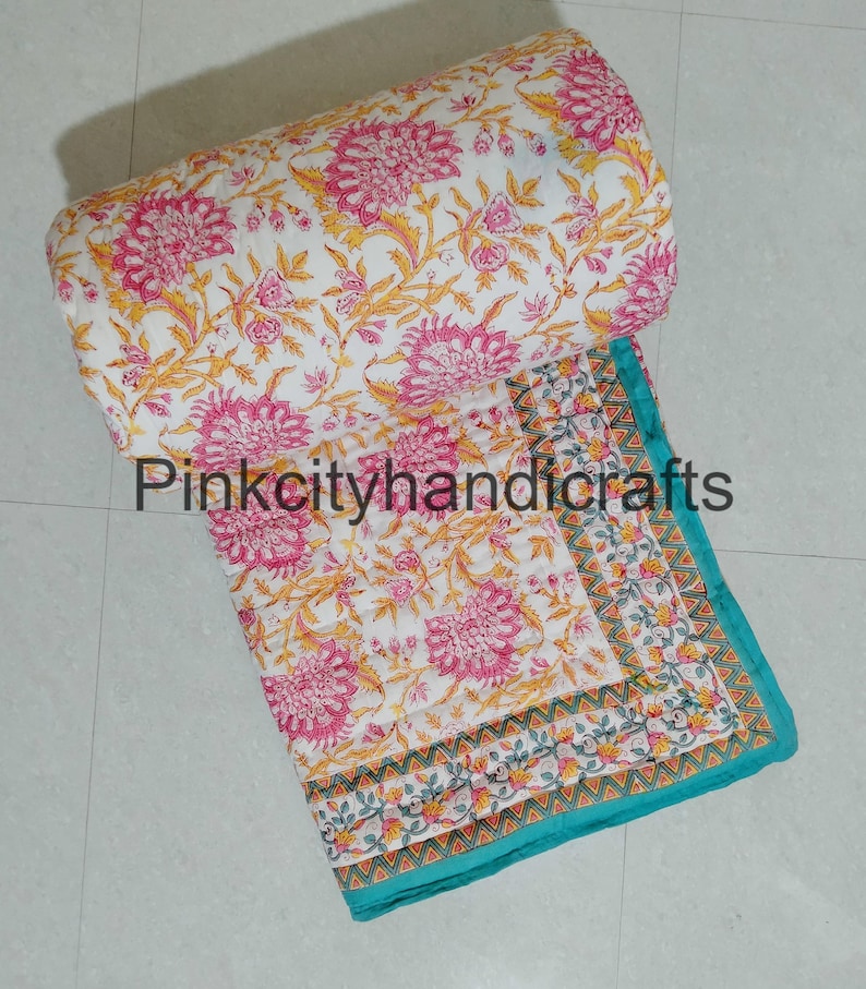 Puede incluir: Una colcha de algod&oacute;n blanco con estampado floral en tonos rosa y amarillo. La colcha tiene un borde turquesa con un patr&oacute;n geom&eacute;trico. La colcha est&aacute; enrollada y el texto "Pinkcityhandicrafts" es visible.