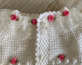May include: White knitted baby cardigan with a lace trim and pink flower embellishments.