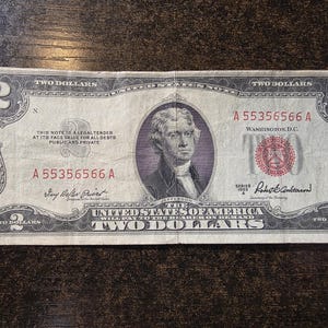 May include: A vintage two-dollar bill featuring a portrait of Thomas Jefferson. The bill is primarily green and gray, with red seals and serial numbers. Text includes "The United States of America" and "Two Dollars."