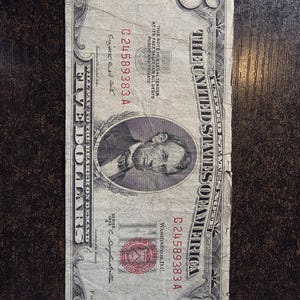 May include: A vintage five-dollar bill from the United States of America. The bill features a portrait of Abraham Lincoln and the text "The United States of America." The serial number is C24589383A. The bill is slightly worn.