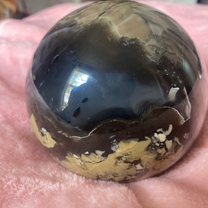 May include: A polished black and brown sphere with white and gold flecks. The sphere is made of natural stone and has a smooth, polished surface.