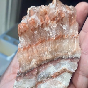 May include: A raw, natural crystal formation with layers of white, clear, and reddish-brown hues. The mineral specimen has a rough, textured surface and a crystalline structure. This unique piece is ideal for collectors or those interested in geology.
