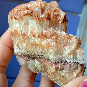 May include: A raw, natural crystal formation with a layered appearance. The crystal features bands of translucent white, orange, and reddish-brown hues. The rough texture and irregular shape highlight its organic origin. The crystal is held in a hand.
