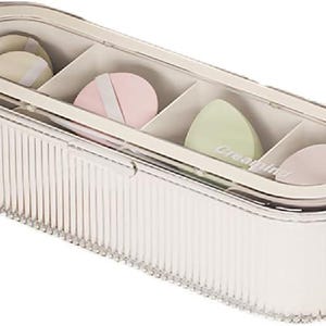May include: A clear, oval-shaped cosmetic organizer with a ribbed design. The organizer contains four compartments, each holding a different colored makeup sponge. The sponges are in shades of beige, pink, and green. The text "Creating" is visible on one sponge.