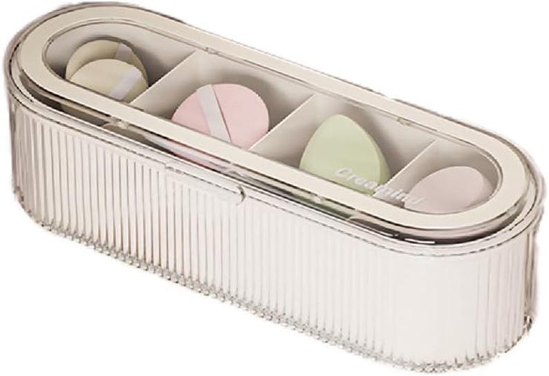 May include: A clear, oval-shaped cosmetic organizer with a ribbed design. The organizer contains four compartments, each holding a different colored makeup sponge. The sponges are in shades of beige, pink, and green. The text "Creating" is visible on one sponge.