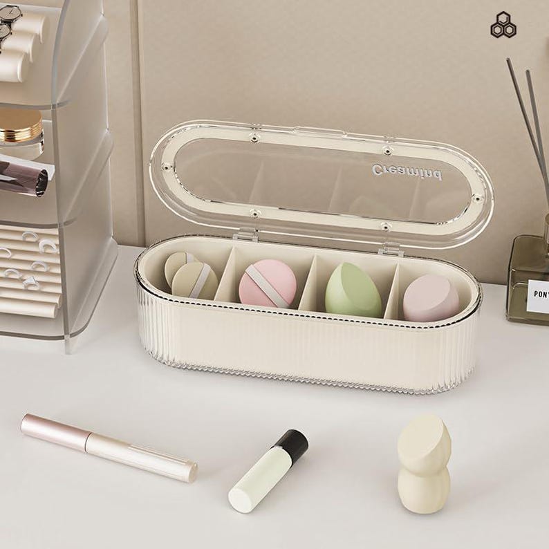 May include: A cream-colored, oval-shaped makeup sponge organizer with a clear lid. The organizer contains several colorful makeup sponges. A pink and white makeup applicator, a white and pink makeup applicator, a green makeup applicator, and a pink makeup applicator are inside the organizer.