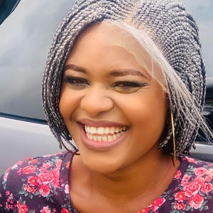 May include: A person smiling, wearing a grey braided wig with a lace front. They are wearing a dark purple top with a floral pattern and gold hoop earrings. The background is a blurred outdoor scene.