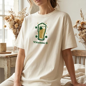 May include: Cream-colored t-shirt featuring a green and gold graphic of a beer mug with shamrocks and the text "Vitamin G". The shirt is made of a soft material and is suitable for casual wear.