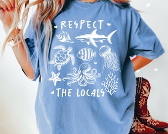 May include: A light blue t-shirt with a white graphic design featuring a shark, turtle, octopus, starfish, coral, and jellyfish. The text "Respect the Locals" is printed on the shirt.