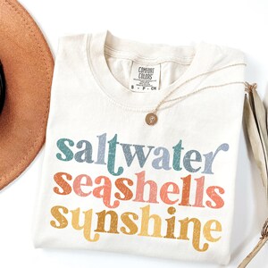 May include: A white t-shirt with the text "saltwater seashells sunshine" in colorful, retro-style lettering. The shirt is laid flat on a white surface with a brown hat and a dried palm leaf.