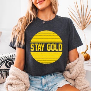 May include: A dark grey t-shirt with a yellow sun graphic and the text "STAY GOLD" printed on it.