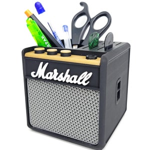 May include: Black Marshall-branded desktop organizer shaped like a guitar amplifier with a black and gray speaker grille. The organizer has slots for pens, pencils, and scissors. It also has a compartment for guitar picks.