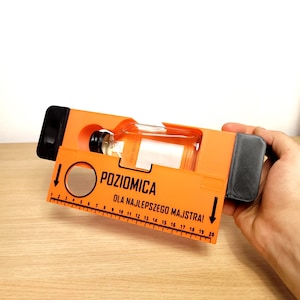 May include: An orange level-shaped gift box with a clear glass bottle inside. The box has black accents and a ruler design with the text "POZIOMICA DLA NAJLEPSZEGO MAJSTRA!" printed on it. The bottle has a black cap.