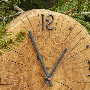 May include: A rustic wooden clock with a natural wood grain finish. The clock face features black metal numerals and hands, with the number 12 prominently displayed. The clock is set against a backdrop of green pine needles.