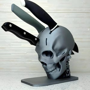 Puede incluir: Un soporte para cuchillos de color gris en forma de cráneo con una base negra. Cuatro cuchillos están insertados en el cráneo, con los mangos sobresaliendo. El cráneo tiene una cara detallada con una nariz y una mandíbula prominentes.