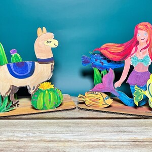 May include: A hand-painted cardboard cutout of a llama standing in a desert landscape with cacti. A second cutout of a mermaid with red hair and a purple tail is surrounded by yellow and blue fish.